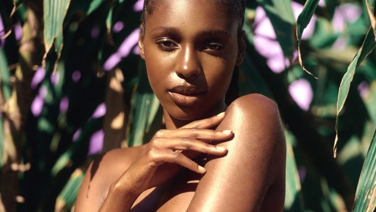 Femme à la peau noire lumineuse touchant son épaule dans un décor tropical — texture légère, zéro film gras, soin pour climat chaud