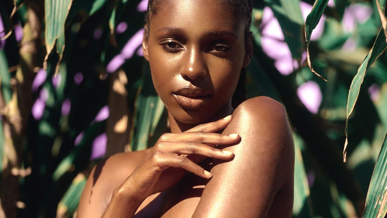 Femme à la peau noire lumineuse touchant son épaule dans un décor tropical — texture légère, zéro film gras, soin pour climat chaud
